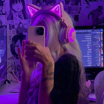 Aesthetic purple-lit photo of a person with blonde hair wearing pink cat-ear headphones, taking a mirror selfie with a phone in front of a computer setup.