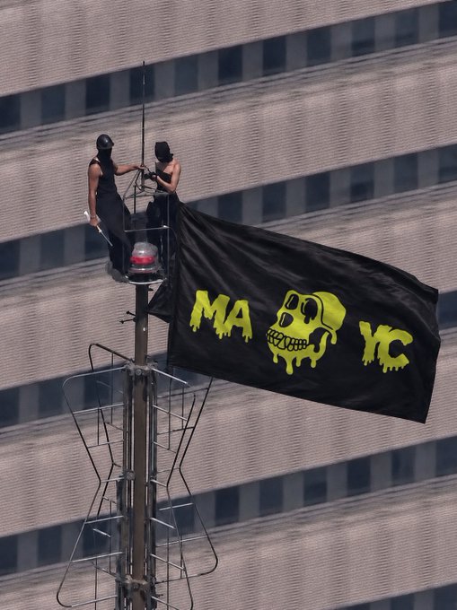 Two people standing on top of a tall communications tower or antenna displaying a black flag with neon green graffiti-style text reading 'MA' and 'YC' flanking a skull design.