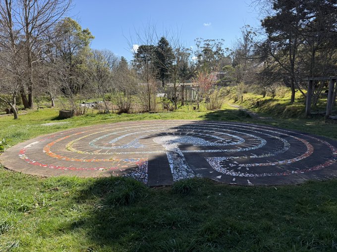 An outdoor circular labyrinth mosaic with colorful concentric rings on a grassy area surrounded by trees and shrubs on a sunny day.