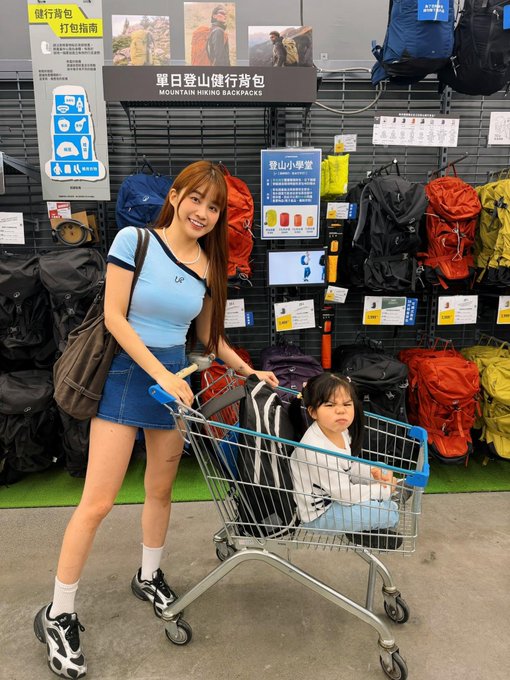 A woman in a blue crop top and denim skirt poses with a child sitting in a shopping cart inside a sporting goods store, with signs reading "MOUNTAIN HIKING BACKPACKS" and Chinese characters displayed on the wall behind them.