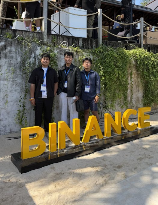 Three young men stand behind large yellow 'BINANCE' letters at what appears to be an outdoor event, with greenery growing on the stone wall behind them.