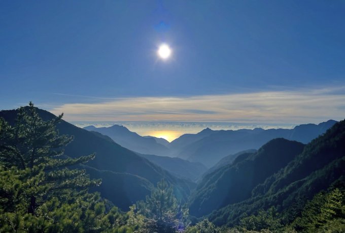 A breathtaking landscape captures the sun shining brightly over a deep mountain valley, with layers of blue-tinted peaks receding into the distance and pine trees in the foreground.