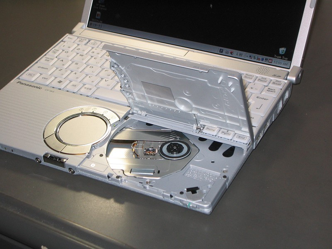 A white Panasonic Let's Note laptop with its optical drive open, revealing the disc tray and internal components on a dark surface.