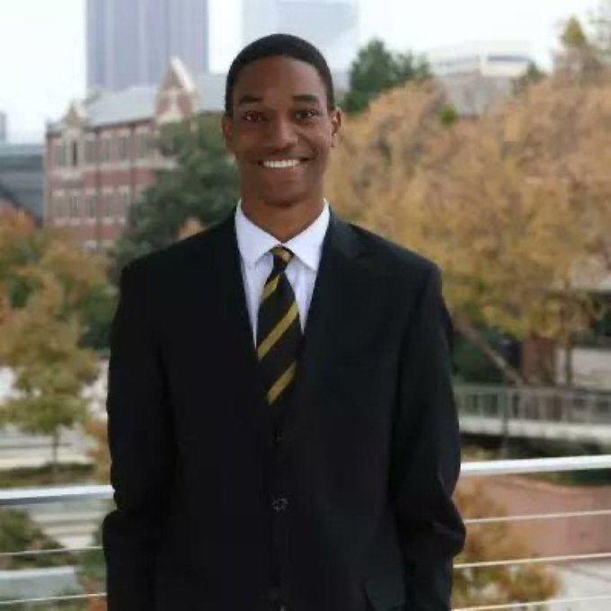 A young man in a black suit with a white shirt and black-and-yellow striped tie smiles at the camera, posing outdoors with autumn trees and city buildings visible in the background.