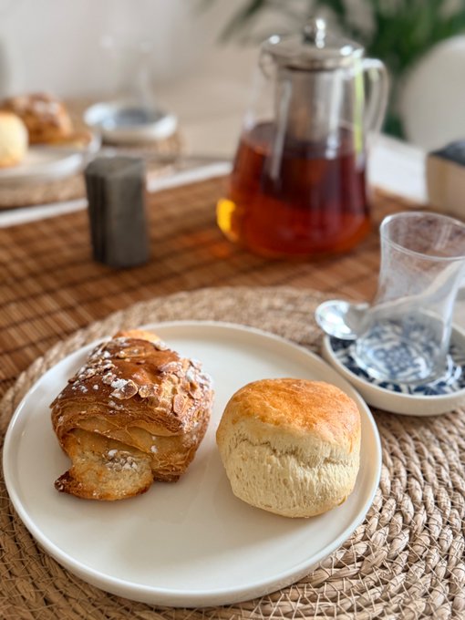 A plate holds a powdered sugar croissant and a plain biscuit, with a glass teapot of tea and an empty glass cup visible on a woven placemat in the background.