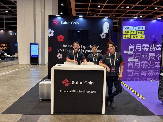 Three staff members stand behind a 'Satori Coin - Physical Bitcoin since 2016' booth at an exhibition, with promotional signage in Chinese including '首月零费率' (first month zero fees).
