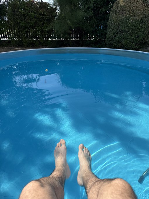 A person relaxes by dipping their feet into a clear blue above-ground pool on a sunny day, with green hedges in the background—capturing the laid-back, "stay hydrated" Sunday vibe of the post.