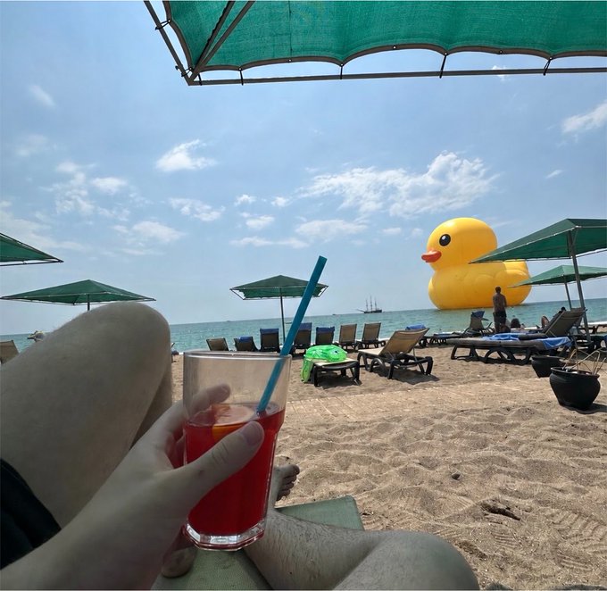 A first-person view from a beach chair holding a red drink with a blue straw, with beach umbrellas and a giant yellow rubber duck visible in the ocean.