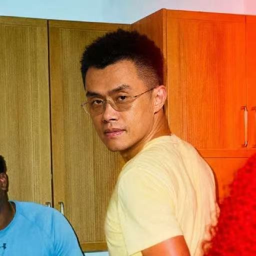 A man with glasses and short dark hair wearing a cream-colored t-shirt looks toward the camera in an indoor setting with wooden cabinets in the background.