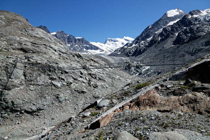 A footbridge spans across a rugged, rocky alpine landscape with a glacier visible in the distance. The bridge, identified as the Corbassière bridge, appears to be a suspension structure crossing a dry riverbed toward the snow-capped peaks beyond.