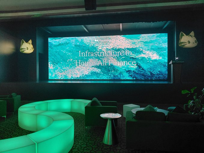 A dimly lit lounge area features a large screen displaying 'Hyperliquid - Infrastructure to House All Finance' over an ocean background, with a glowing green curved sofa and cat logos on the walls.