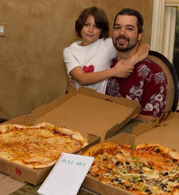 A man and child pose behind two large pizzas in open delivery boxes, one cheese and one topped with olives and vegetables, with a notepad reading "7641 ARC1." The image ties to the post by illustrating a relatable real-world purchase—ordering food for the family—that could conveniently be paid for using cryptocurrency instead of traditional currency.