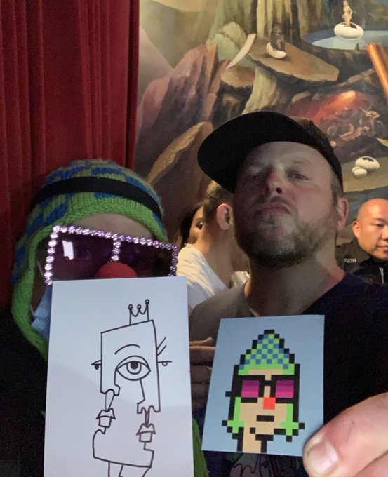 Two men posing while holding up printed character illustrations, with one wearing a green and blue striped beanie and the other in a black cap, set against a mural-covered wall.