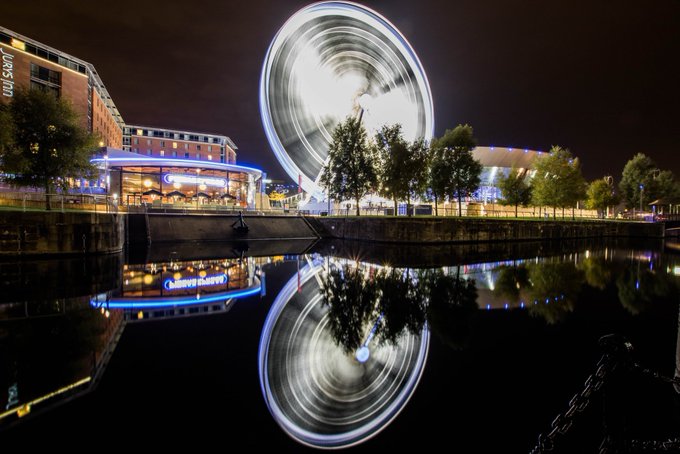 The image captures a cityscape at night featuring a large white Ferris wheel with its lights glowing brightly against the dark sky. A body of water nearby reflects the radiant spectacle above, adding depth and visual interest to the scene. The photograph is taken from the perspective of a pier or dock area, providing an elevated viewpoint of the Ferris wheel and surrounding landscape. 

The image also includes a caption below the photo that discusses the significance ofGN CT @RiverdotInc in relation to the Ferris wheel. This connection adds intrigue to the overall composition while conveying the essence of the city's night-time ambiance, highlighting the theme of investing in the city as seen through the Ferris wheel and how it can contribute positively to its growth.
