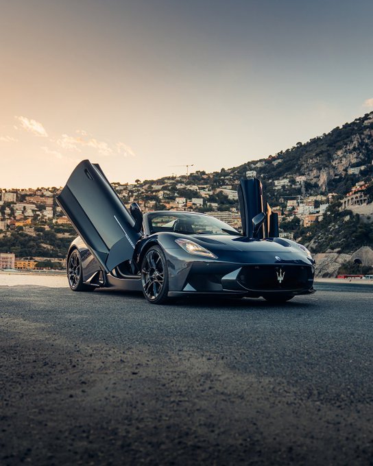 The image features a blue sports car with its doors open, parked on a road near some buildings. The car appears to be an expensive luxury vehicle, possibly a sports car or a supercar. The scene takes place at sunrise, creating a serene atmosphere as the sun rises over the horizon and casts a warm glow on the surroundings.