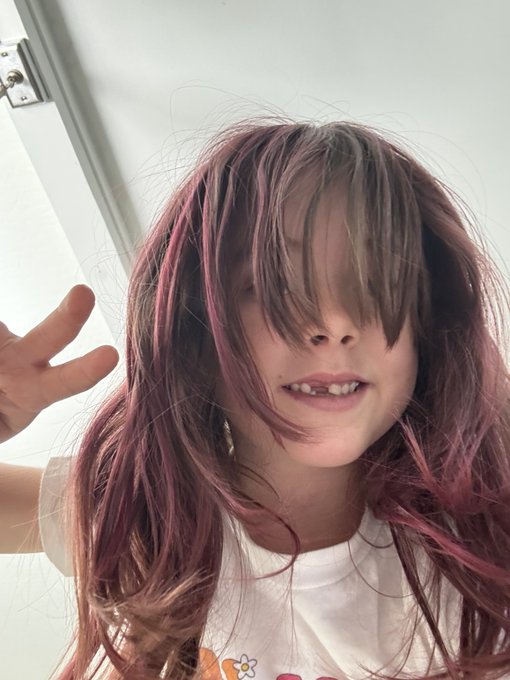 A child with long pink-dyed hair covering their eyes smiles playfully while making a peace sign in front of a white door.
