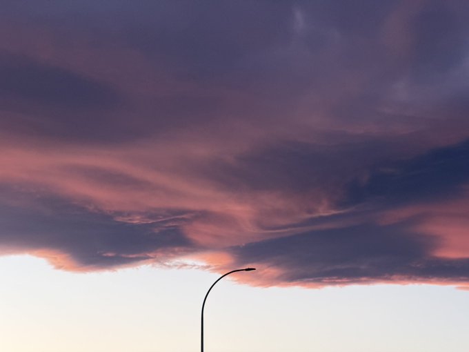 A silhouette of a curved street lamp stands beneath a dramatic sky filled with pink and purple clouds at sunset.