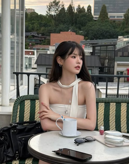 A young woman sits at an outdoor café table with a coffee cup, smartphone, and sunglasses, looking off to the side with a cityscape behind her.