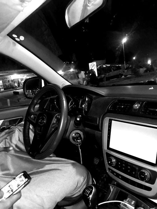 A black-and-white photograph taken from inside a car, showing the dashboard, steering wheel with a Volvo logo, and a person holding a smartphone, with a gas station visible through the windshield at night.