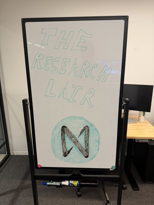 A whiteboard on a stand is labeled 'THE RESEARCH LAIR' in green marker with a drawn 'N' logo inside a teal circle, set up in an office environment.