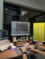 A photo of a person sitting at a desk with two computer monitors in a dimly lit home office or workspace.