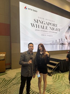 A man and a woman pose together for a photo at a 2026 HTXDAO Singapore Night event, standing in front of a large projection screen displaying '2026 HTXDAO SINGAPORE NIGHT 2026 HTXDAO VIP Night' with the date '2026.11 Singapore' and a city skyline image.