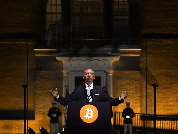 A man in a dark suit stands at a podium displaying the Bitcoin logo, gesturing with open arms during what appears to be a speech or presentation at night, with military personnel standing guard behind transparent barriers.