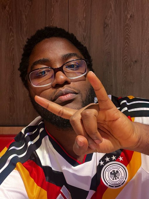 A man wearing glasses and a Germany football jersey with the DFB logo making a peace sign or gun gesture with his fingers.