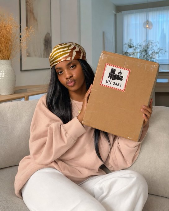 A woman wearing a patterned headscarf and pink sweatshirt sitting on a beige couch, playfully holding a cardboard shipping box marked with a fragile label and 'UN 3481' (lithium battery warning).