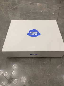 The image shows a white box with blue text that says "Love HTC" written on it, placed on a gray floor. The box appears to be new and unused, possibly indicating that it has been recently purchased or received as a gift. The photo is taken from an angle, which adds depth to the scene and emphasizes the size of the box.