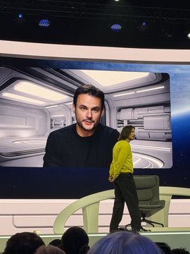A man in a black jacket stands on a stage with a large screen behind him displaying a close-up image of his own face.
