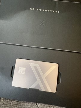 A sleek black card with a large "X" logo sits on a box labeled "TAP INTO EVERYTHING," teasing the upcoming launch of X Money. The minimalist design hints at the new digital payment feature Elon Musk's platform is rolling out.