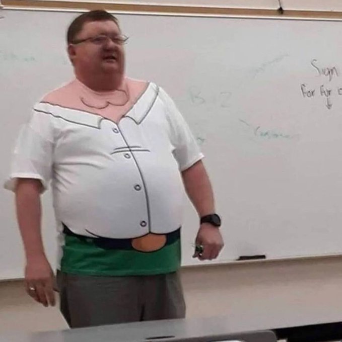 A man wearing glasses stands in front of a whiteboard, humorously wearing an inflatable costume that gives him an exaggerated large belly with drawn-on shirt buttons and a green belt/sash design.
