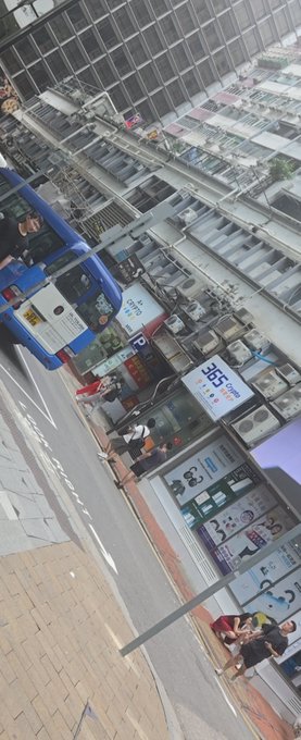 The image shows a busy street scene with people walking along the sidewalk next to tall buildings. There are two buses parked near each other, one blue and one white. The perspective of the photo suggests that it was taken from someone standing on the sidewalk or possibly in front of one of the buildings. A person is also seen carrying a suitcase, indicating they might be traveling or moving between locations.