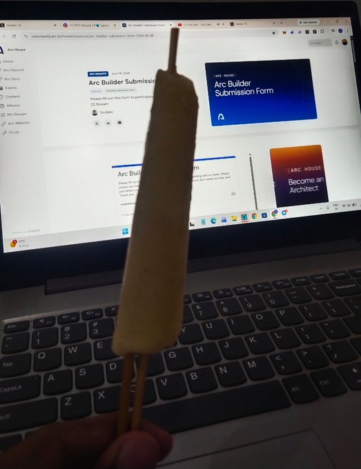 A photo of a hand holding a corn dog skewer in front of a laptop screen displaying an 'Arc Builder Submission Form' page on community.arc.