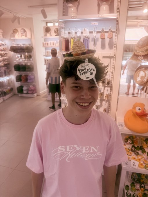 A smiling young man in a pink 'SEVEN NINE' t-shirt wearing a novelty sombrero with a tag reading 'Stay Positive' in a brightly lit store, with a rubber duck visible on the right.