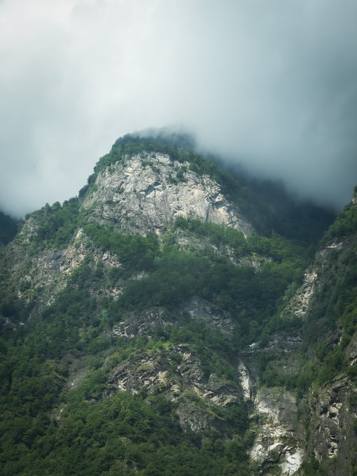 A misty, dramatic mountain peak with rocky cliffs covered in lush green vegetation, partially obscured by low-hanging clouds.