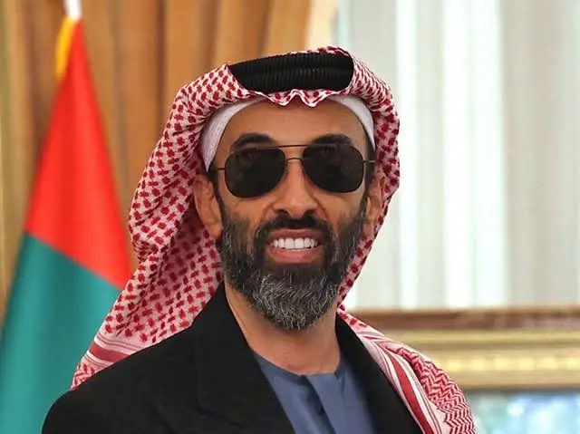 A portrait of a smiling man with a beard wearing sunglasses and a traditional red and white checkered keffiyeh, with a red, white, and green flag visible behind him.
