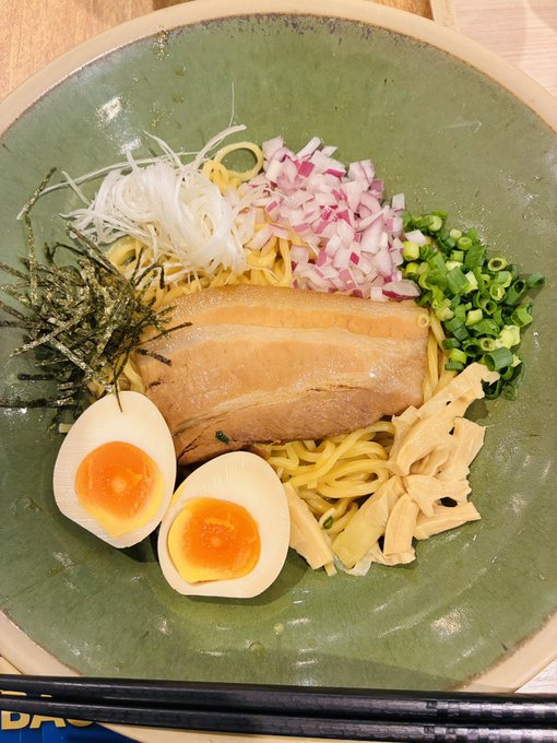 A green ceramic bowl filled with ramen, featuring a soft-boiled egg cut in half with vibrant orange yolks, chashu pork, and topped with shredded radish, diced red onions, green onions, nori strips, and bamboo shoots, with black chopsticks resting beside it. The Japanese post expresses frustration with Bitradex and shares a photo of the ramen the poster ate earlier, tying the meal to their late-night venting.
