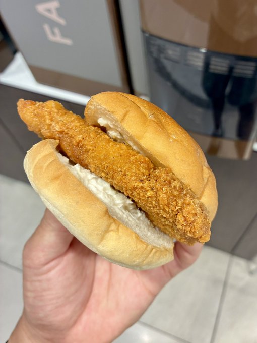 A hand holding a fried fish sandwich on a soft white bun, with a Longchamp or similar shopping bag visible in the background.