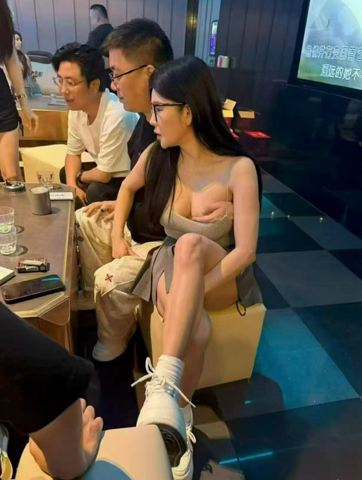 The image shows a group of people sitting around a table at a restaurant or bar. A woman with long hair wearing glasses sits next to a man holding a baby while another person holds a cup, possibly enjoying their time together. The background features a TV screen mounted on the wall and other people seated in the room.