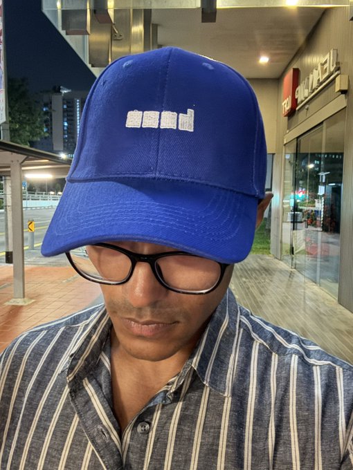 A person wearing black-rimmed glasses and a blue "raid" baseball cap, with the brim pulled down to obscure their eyes, stands outside at night near a Subway restaurant, illustrating the post about giving away "raid" branded hats.