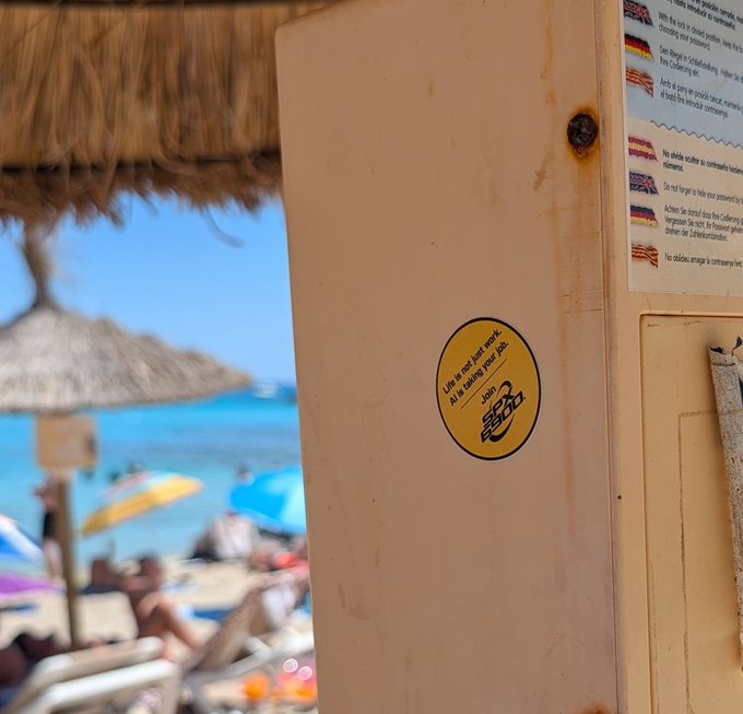 The user wants a caption describing the image.
I need to identify key elements:
- Foreground: A beige utility box or cabinet.
- On the box: A yellow circular sticker.
- Text on sticker: "Life is not just work. AI is taking your job." and a logo "SPX6900".
- Background: A blurred beach scene with blue water, sand, umbrellas, and people.
- Right side of the box: A white paper with text in multiple languages (English, German, Spanish, French, Italian, Portuguese) warning about robots taking jobs ("No robots working on construction sites", "Do not forget to take your personal belongings", etc.).

Synthesizing the description:
The image shows a close-up of a beige utility box in the foreground, featuring a yellow sticker that reads "Life is not just work. AI is taking your job" with an "SPX6900" logo. In the background, out