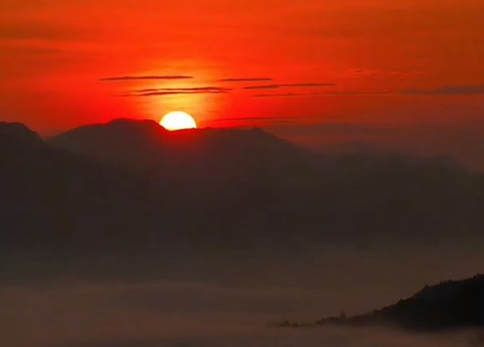 A stunning red and orange sunset with the sun dipping behind silhouetted mountains, casting a warm glow over misty valleys.