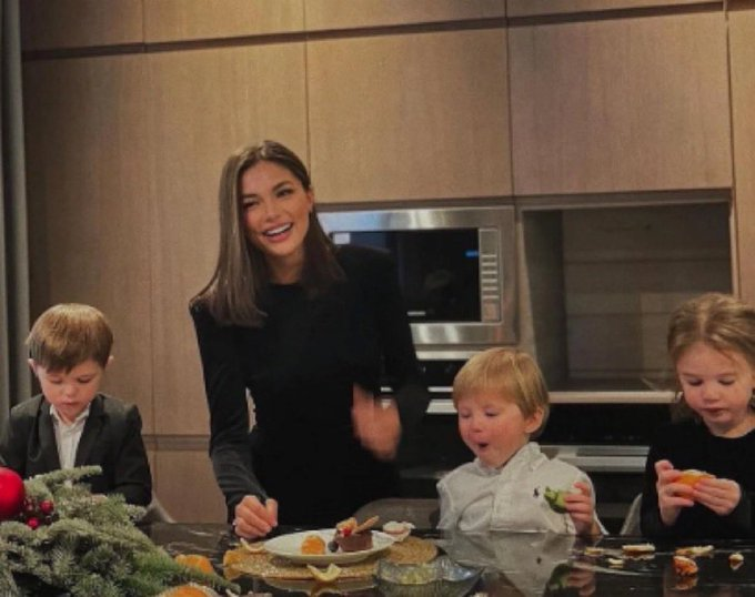 A smiling mother stands at a modern kitchen island with three young children gathered around plates of food and citrus garnishes. The joke plays on the meme-worthy image of a wealthy-looking family in a sleek, upscale kitchen, implying their fortune came from the father's relentless memecoin speculation rather than legitimate work.