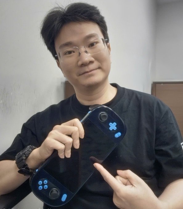 An Asian man wearing glasses and a black t-shirt smiles while holding up a handheld gaming device featuring dual screens, joystick, and buttons.