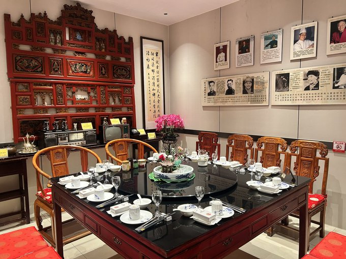 An elegant traditional Chinese dining room featuring a dark wooden table set with dishes, ornate red wooden chairs, a decorative carved wooden screen, and a wall displaying framed portraits and Chinese calligraphy.