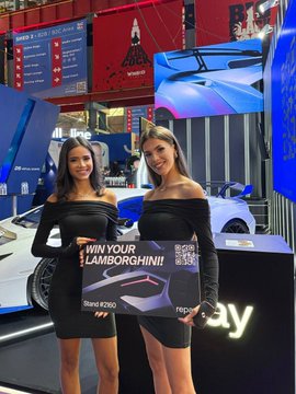 Two women in black off-shoulder dresses holding a sign that reads 'WIN YOUR LAMBORGHINI!' at what appears to be a trade show or expo booth.