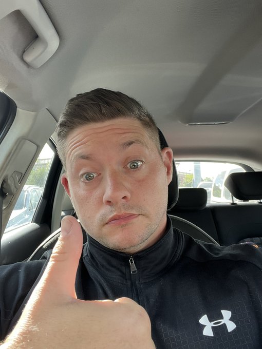 A man wearing an Under Armour jacket sits inside a car and gives a thumbs-up to the camera.