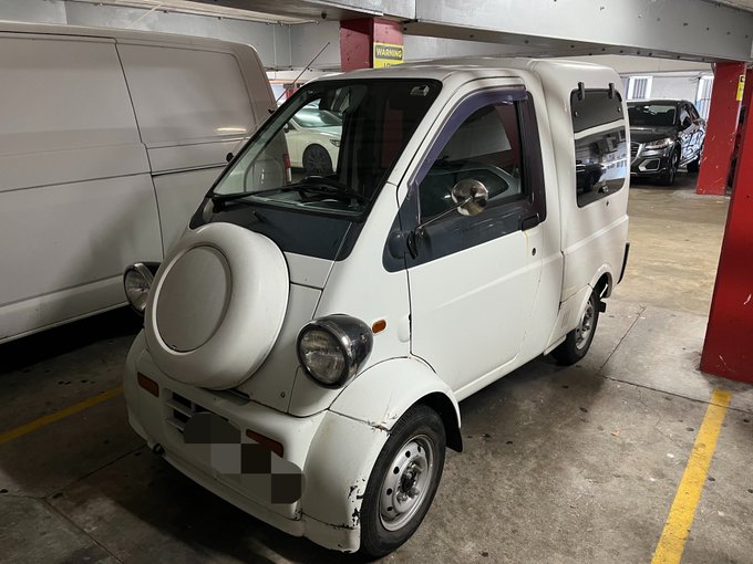 A small white vintage Japanese kei-style van parked in an indoor parking garage, featuring a spare tire mounted on the front hood and a rounded, compact design.